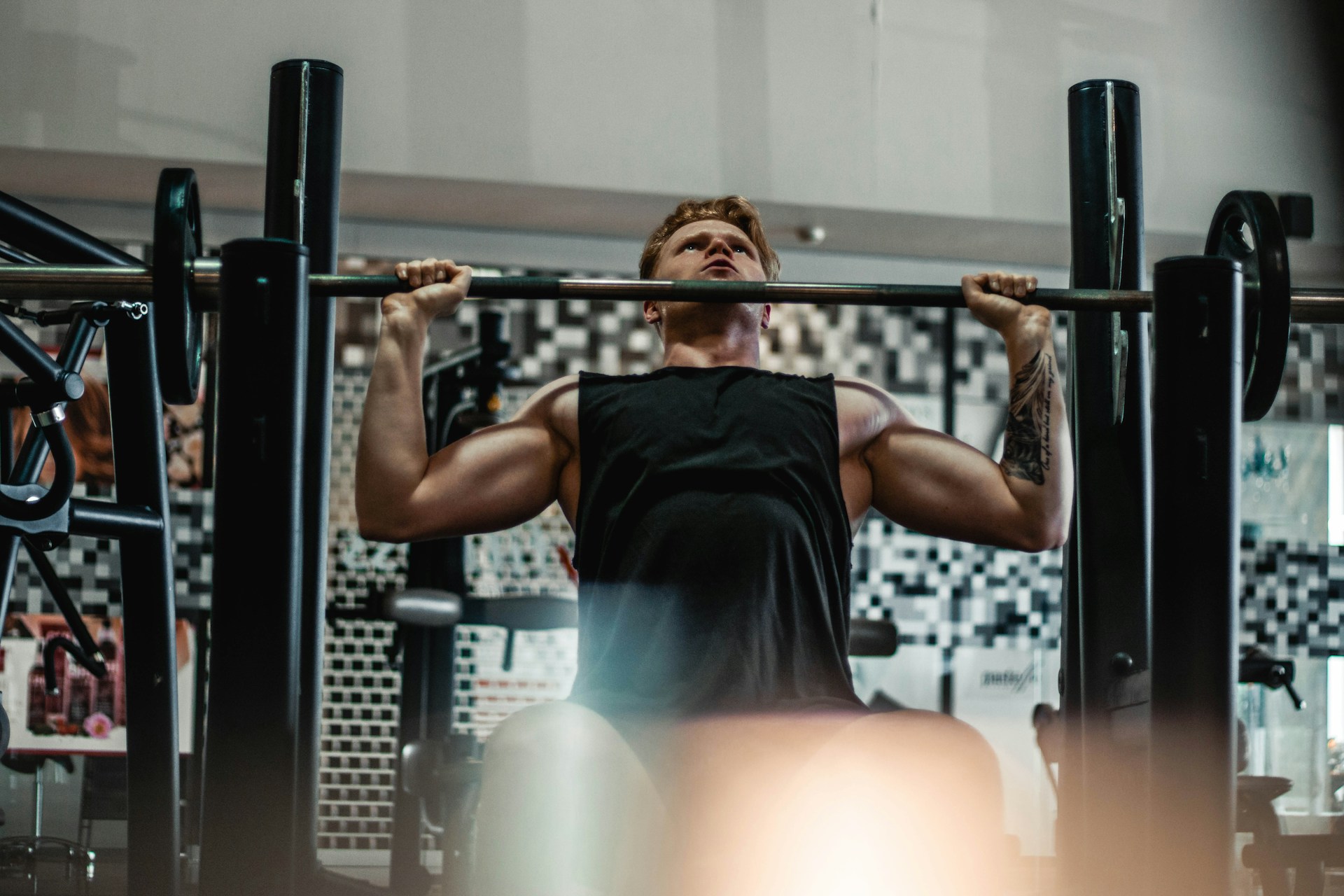 A lifter pressing a barbell overhead at a squat rack in a dimly lit gym